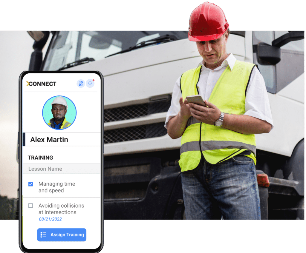 A man in a red hard hat and yellow safety vest uses a smartphone in front of a truck, seamlessly integrating into the driver monitoring system. Next to him, a mobile app screen displays a user profile, showing training details and an option to assign further training.