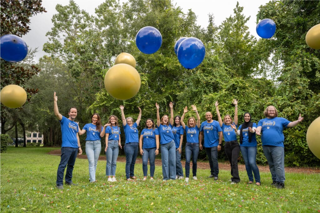 MVR Monitoring team in blue shirts stand on grass, smiling and raising their arms as large yellow and blue balloons float above them. Trees are in the background.