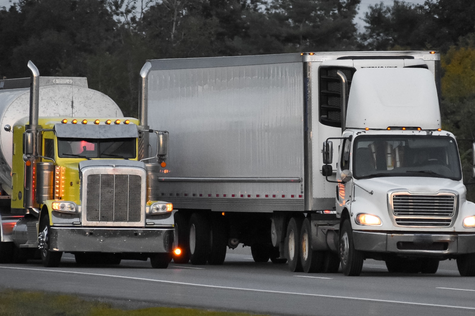 Two semi-trucks are driving side by side on a highway. The one on the left is yellow with a visible silver exhaust stack, and the right one is white, both surrounded by trees.